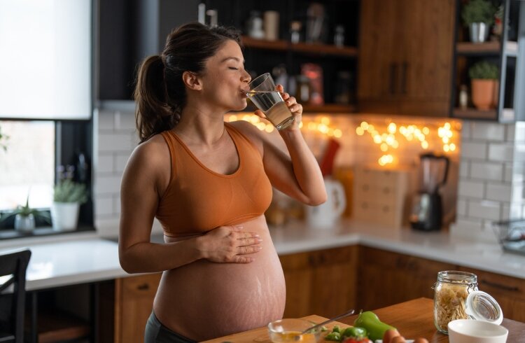 Pregnant woman drinks glass of water while cooking Pregnant woman drinks glass of water